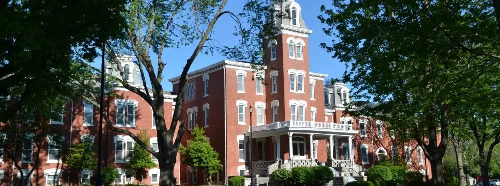 Main Hall on Cottey College's campus is a traditional brick building with white trim, surrounded by lush trees and bushes.