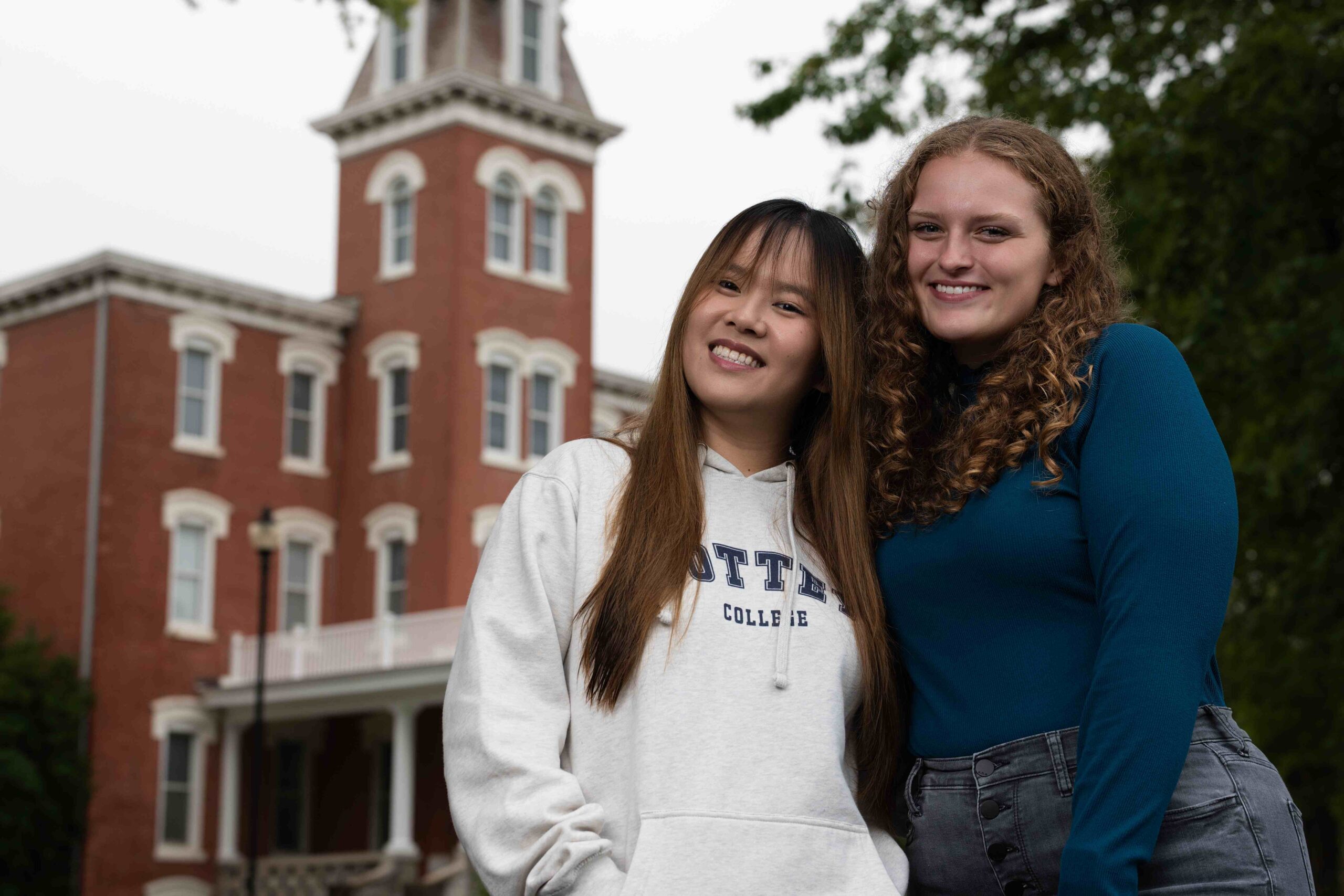 Two college students posing in front of a historic brick building, one is wearing a white sweatshirt and the other a teal long-sleeve shirt