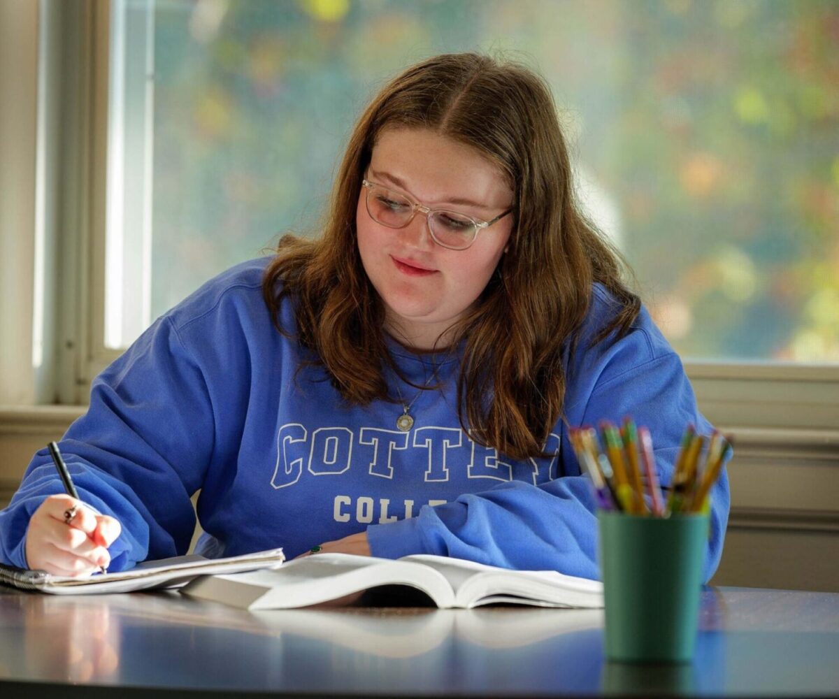 Smiling college student studying, writing in a notebook, and wearing a blue Cottey College sweatshirt.