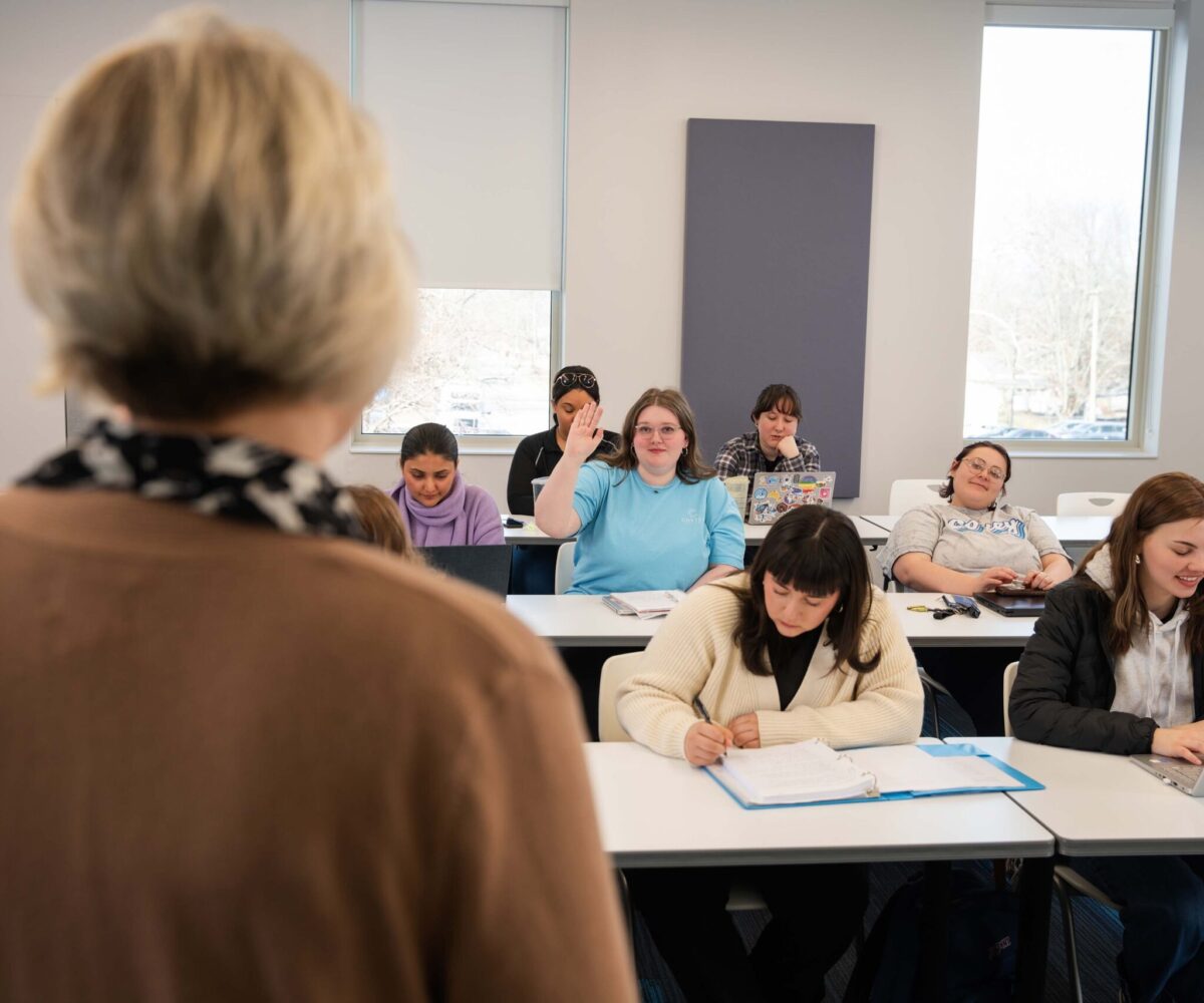 Female students in a classroom, one raising her hand to ask a question.