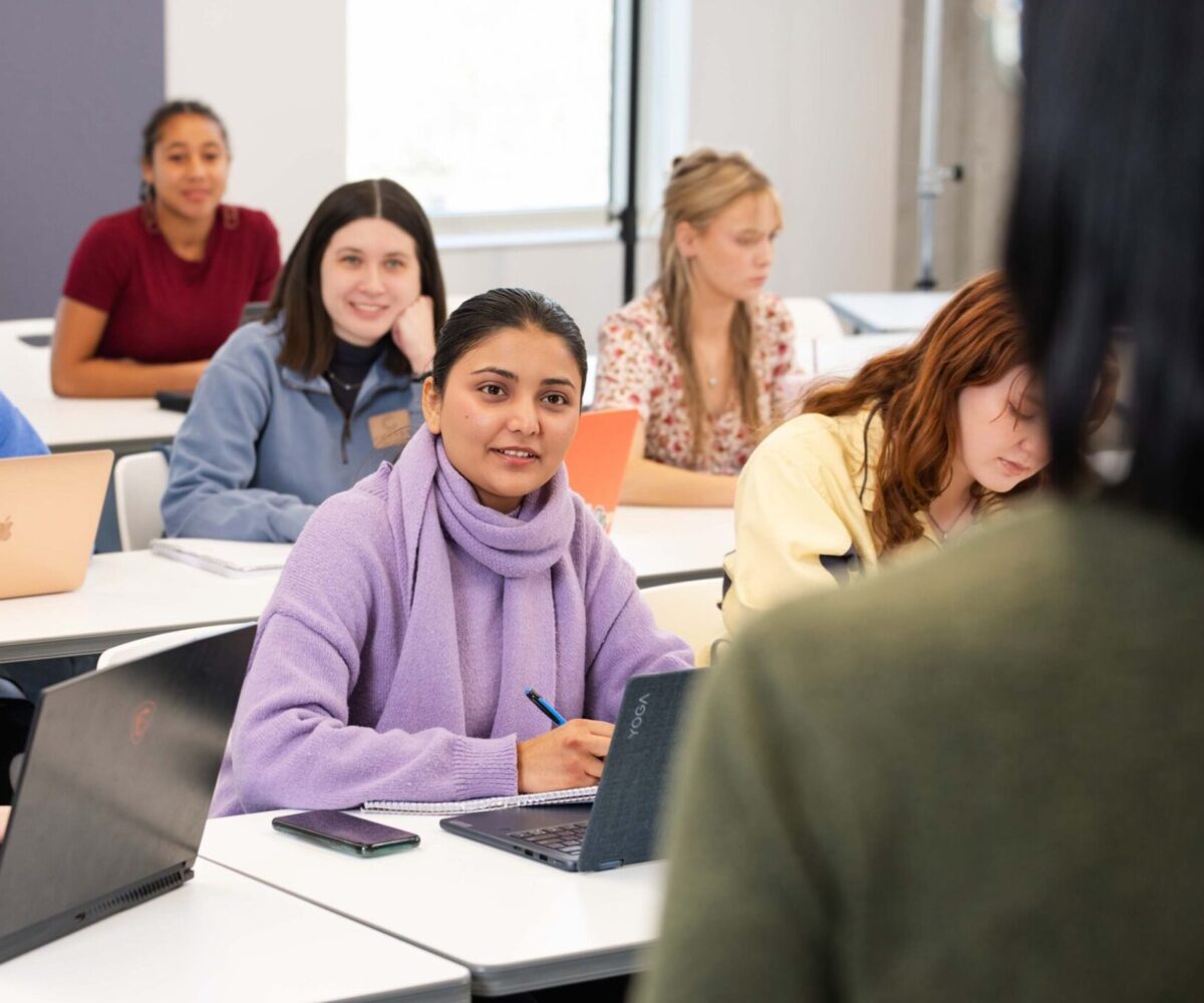 A student in a purple sweater taking notes while looking at her professor.