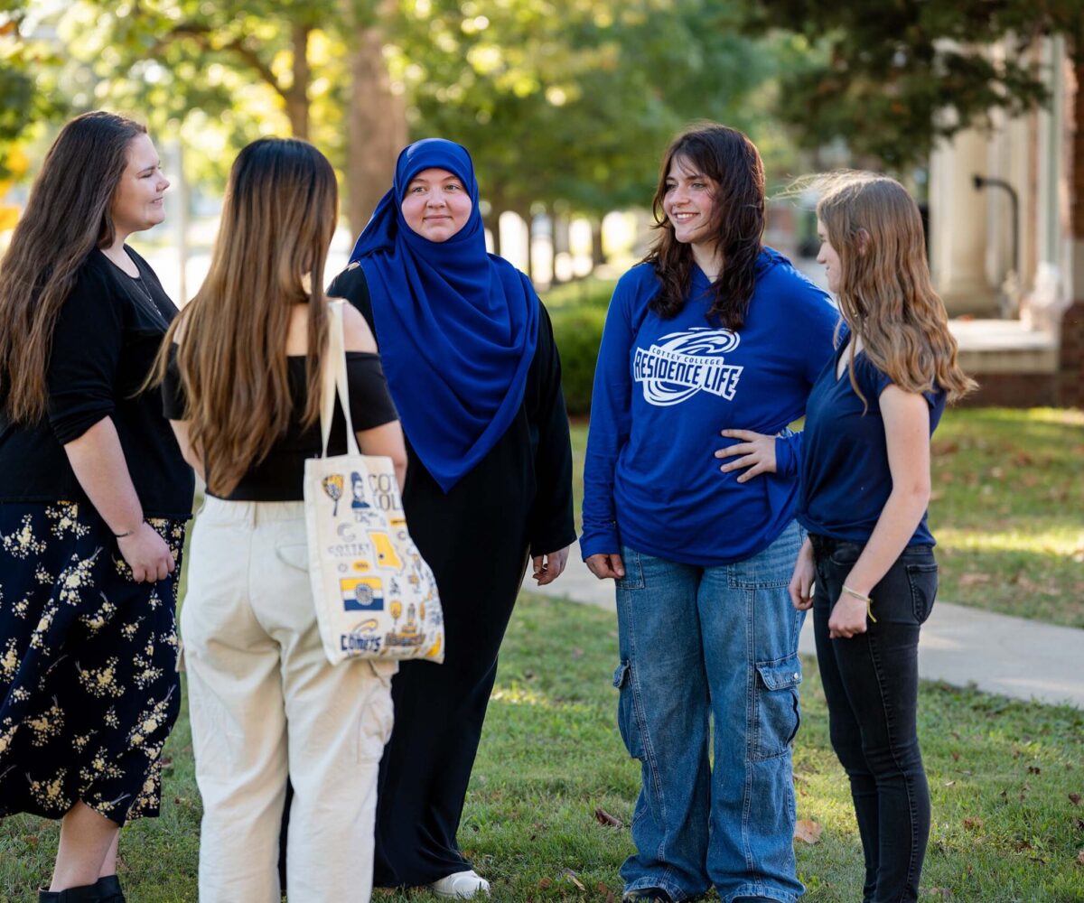 College students standing in a group on a green lawn surrounded by trees.