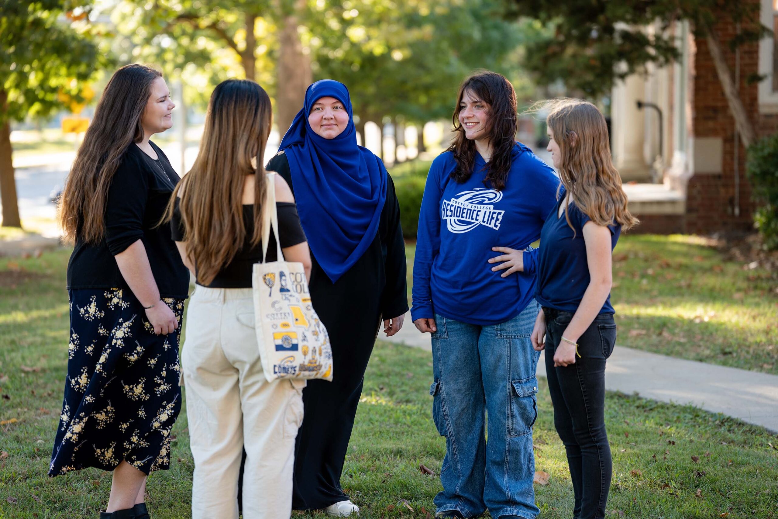 A group of college students standing in a circle talking in a beautiful green lawn with sunlight coming through the trees behind them.