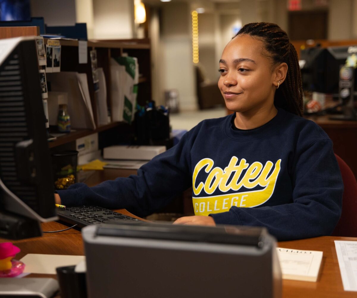 Student working an internship, sitting at a desk, wearing a Cottey College sweatshirt.