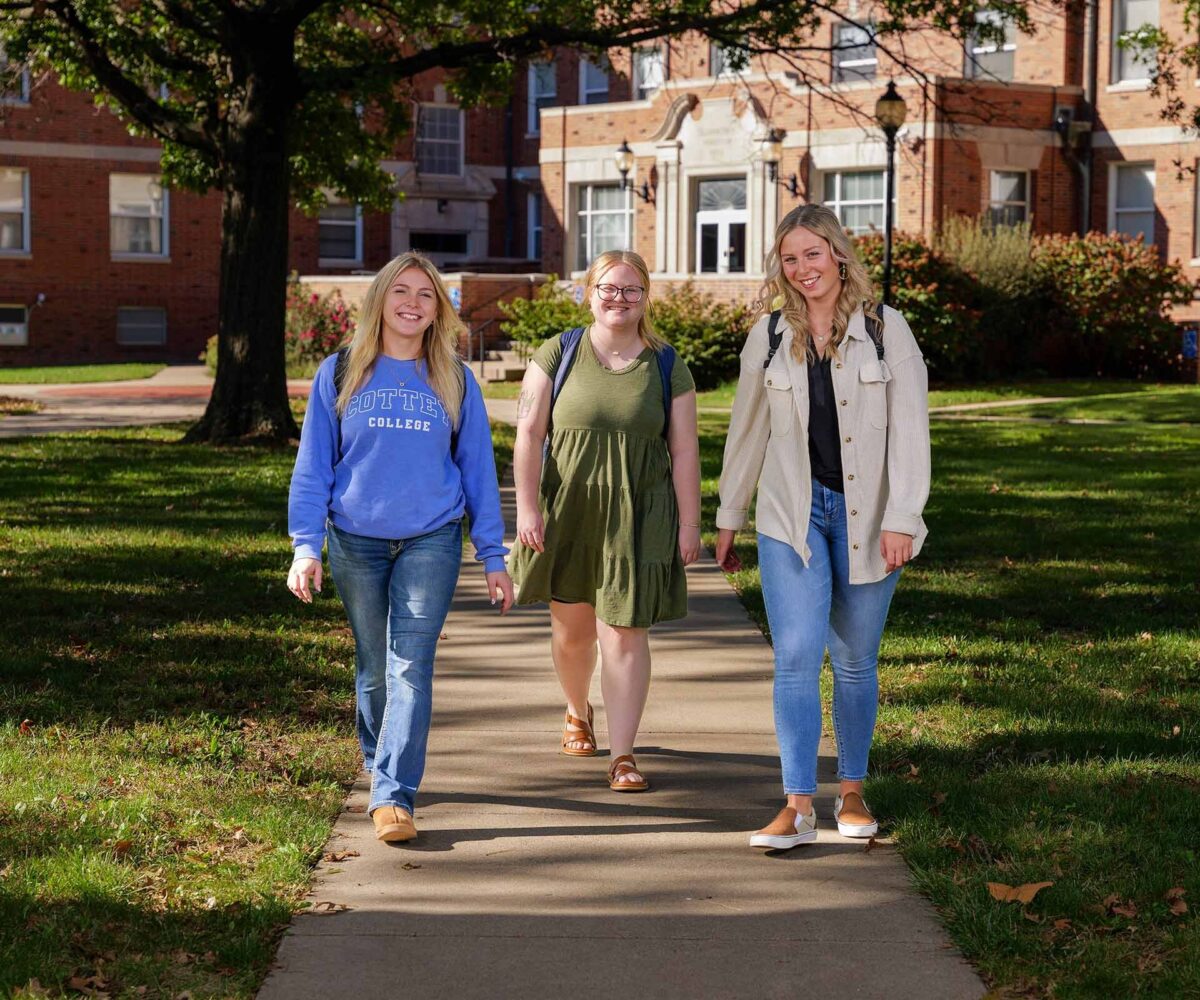 Three female college students walking on a beautiful campus with green grass and historic brick buildings.