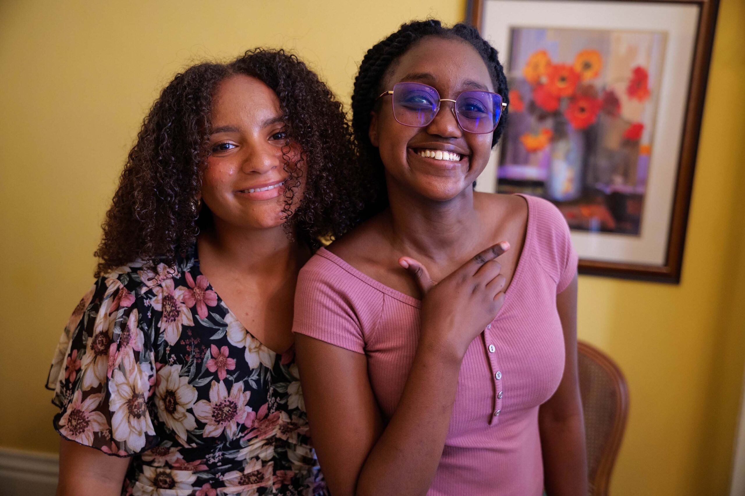Two college students, one wearing a pink top with pink sunglasses, the other a floral shirt, smiling at the camera in front of a yellow wall with a flower painting hung behind the students