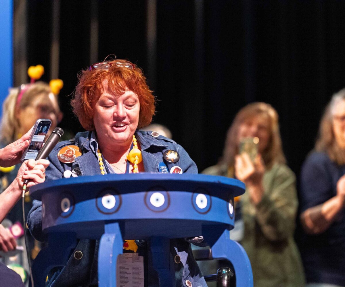 Woman with red hair, wearing jean jacket, talking at podium.