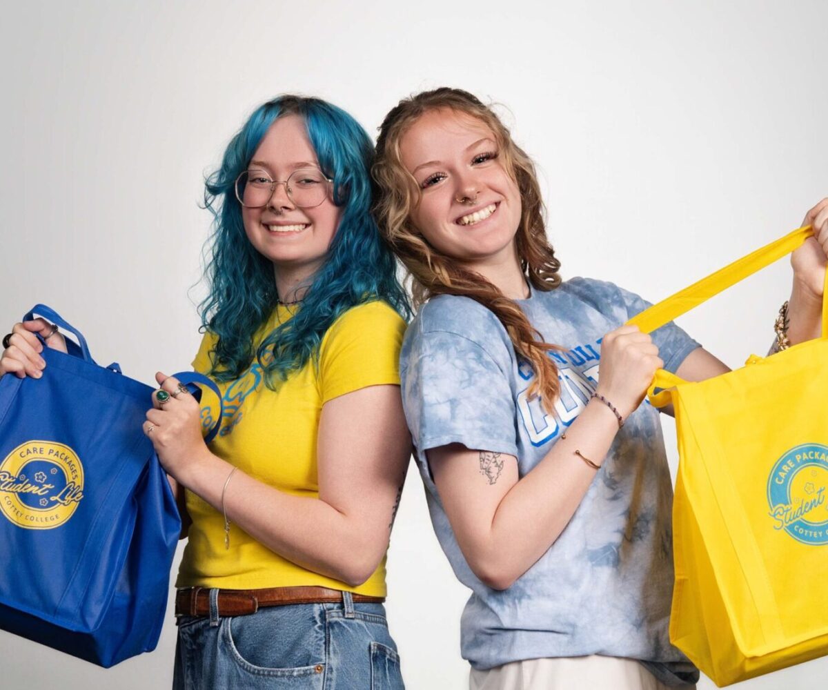 Two Cottey College students standing back-to-back, holding up brightly-colored bags full of treats.
