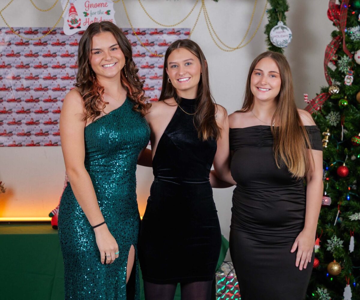 Three students in formal gowns posing in front of a Christmas tree at Cottey College's annual 'Hanging of the Greens' dinner.