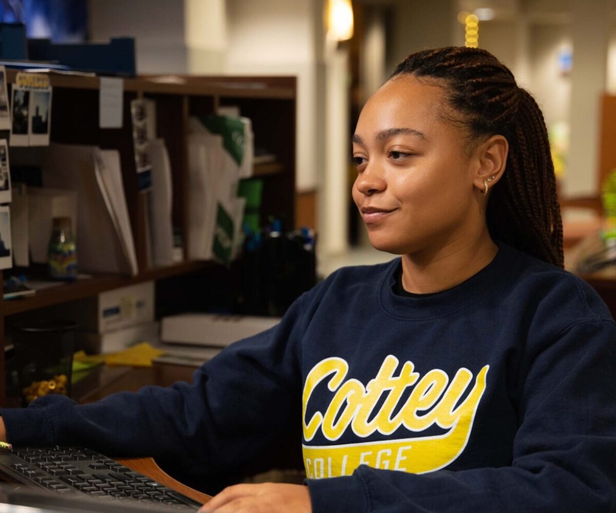 Cottey College student confidently working at a desk in an office, wearing a navy Cottey sweatshirt.