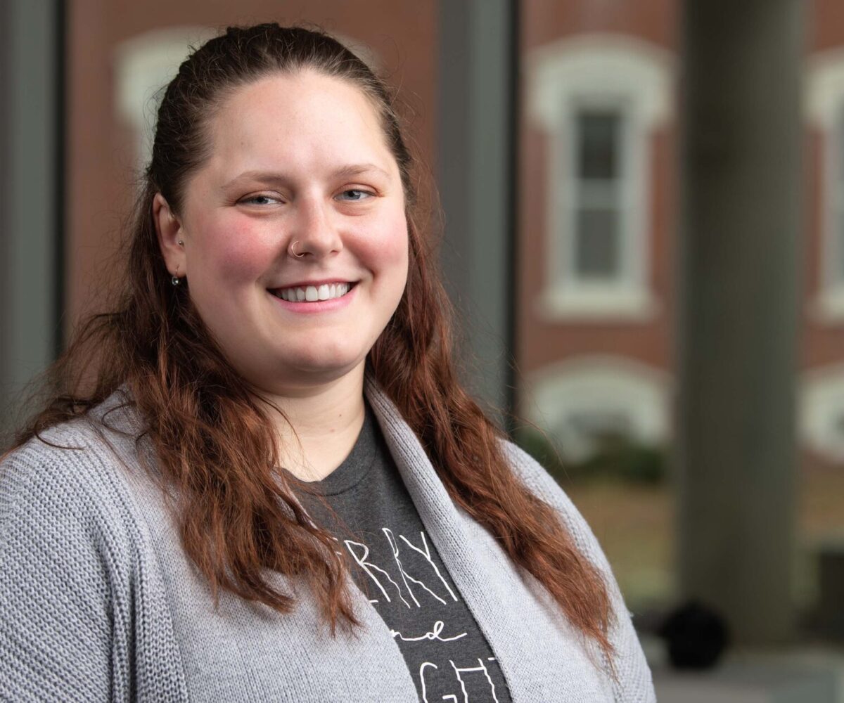 A woman with a gray sweater and a golden hoop nose ring smiles at the camera.
