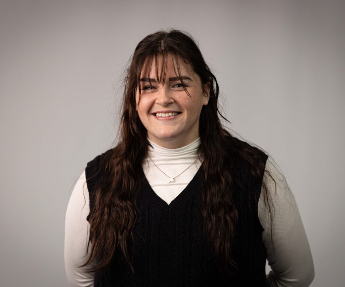 A woman with long, brown hair is wearing a black vest, white turtleneck, and gold necklace.