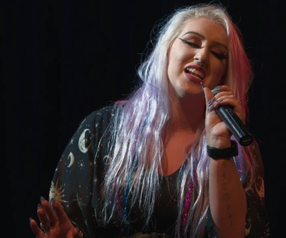 A woman with blond and purple hair singing into a microphone with passion.
