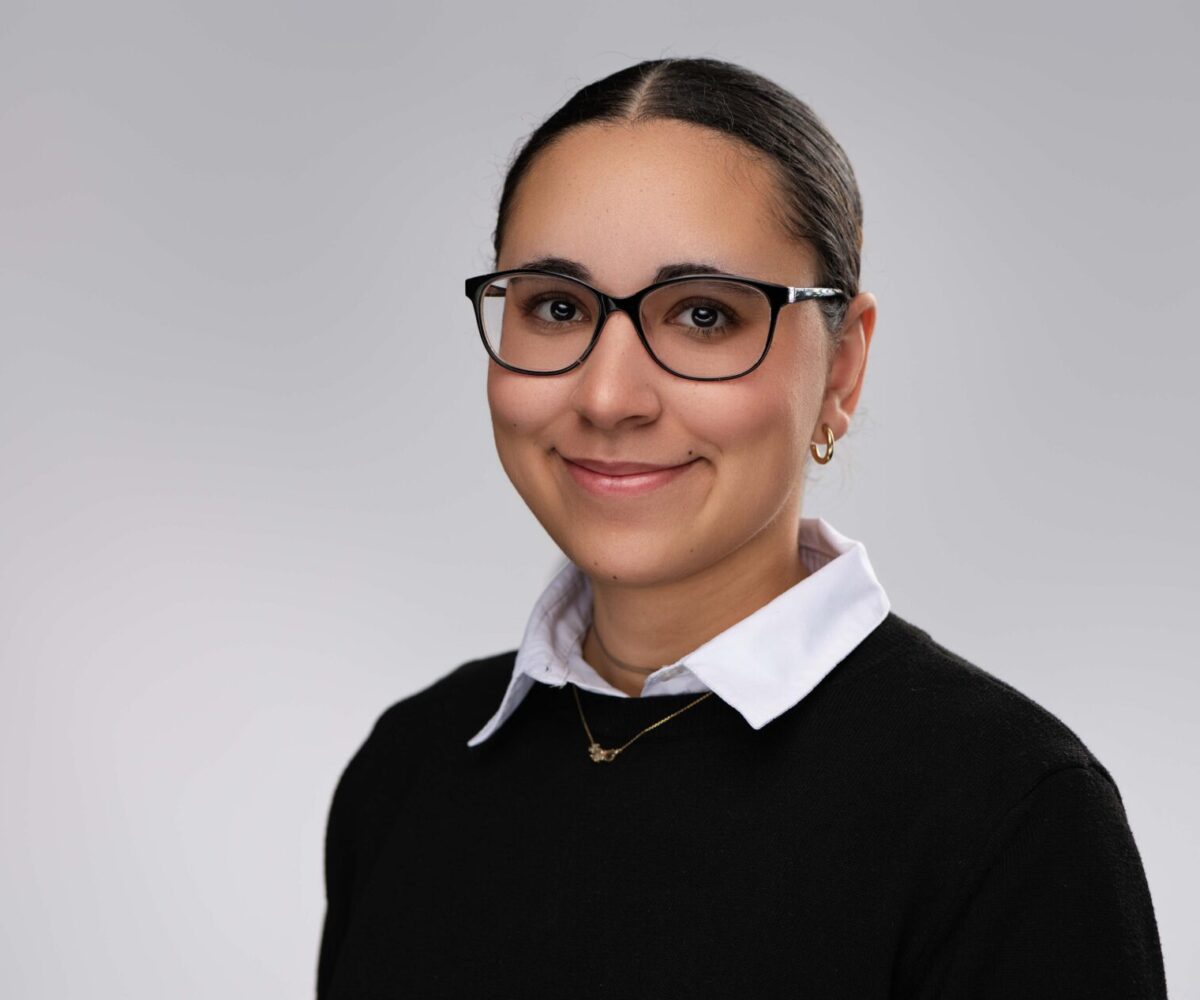 Young woman posing for a picture with black sweater, white collared shirt, and black frame glasses.