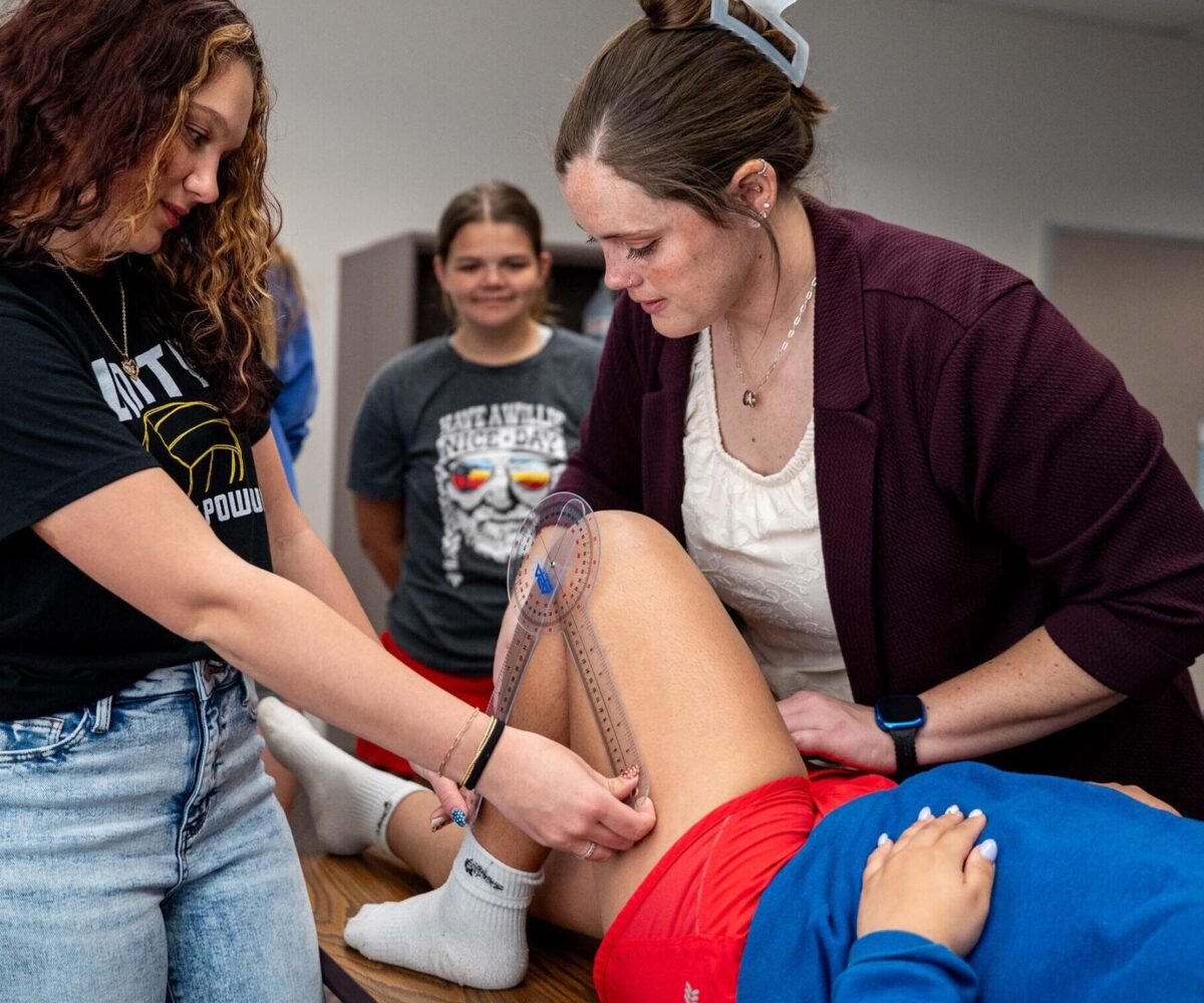 A student uses an instrument to measure the knee of an athlete; her teacher watches over to guide her.
