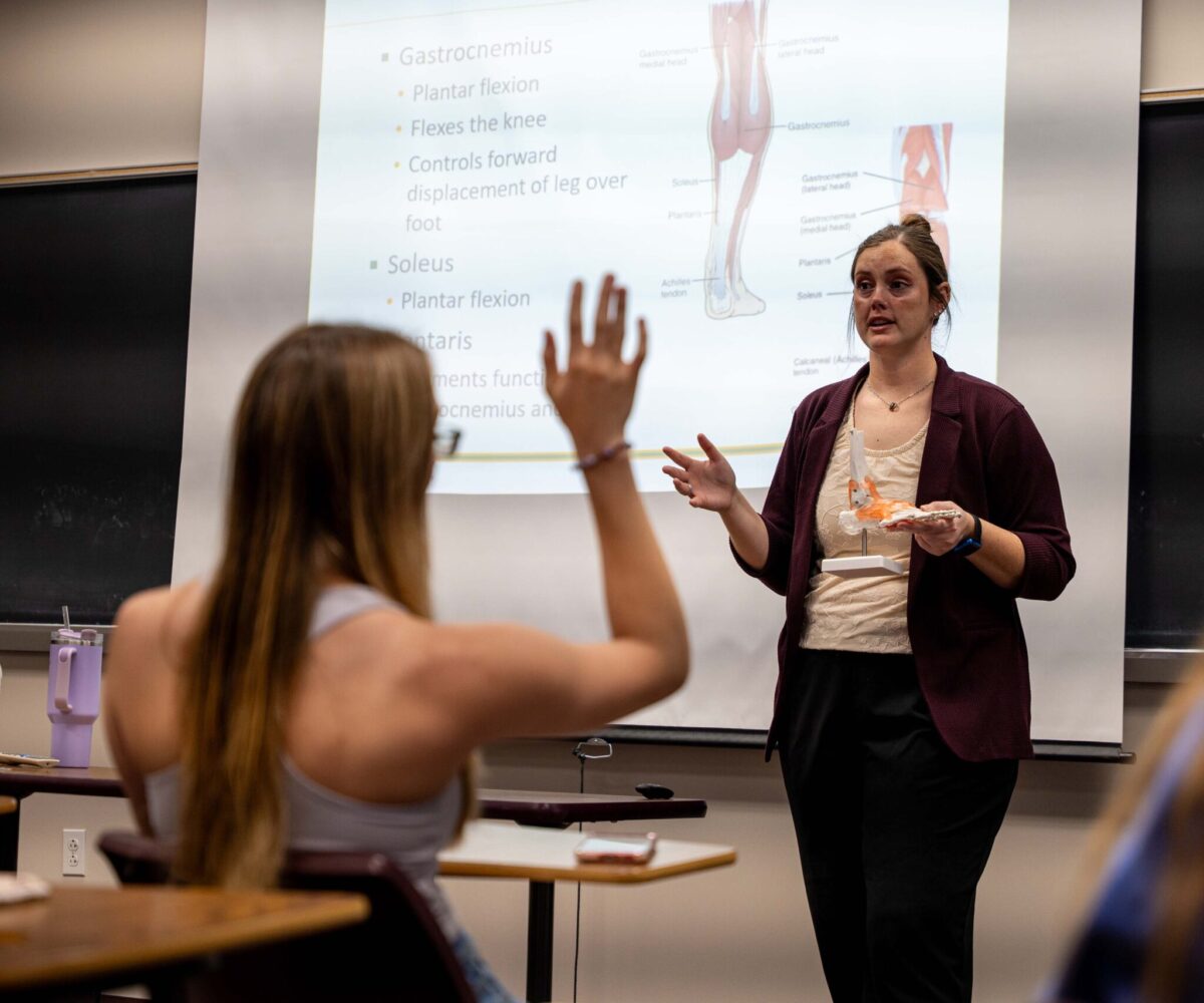 A student raises her hand in a sports science class; an illustration of the musculature of the human leg is projected onto the wall.